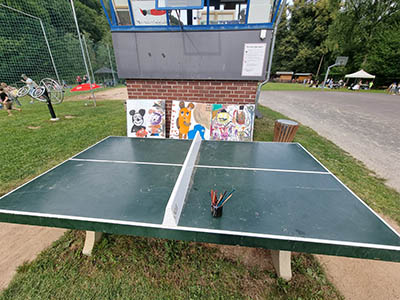 Nach dem Malen; die Leinwände stehen an der Wand hinter der Tischtennisplatte und diese ist bereits geputzt. Es stehen noch ein Glas mit Pinsel auf der Platte. Diese müssen noch gereinigt werden.